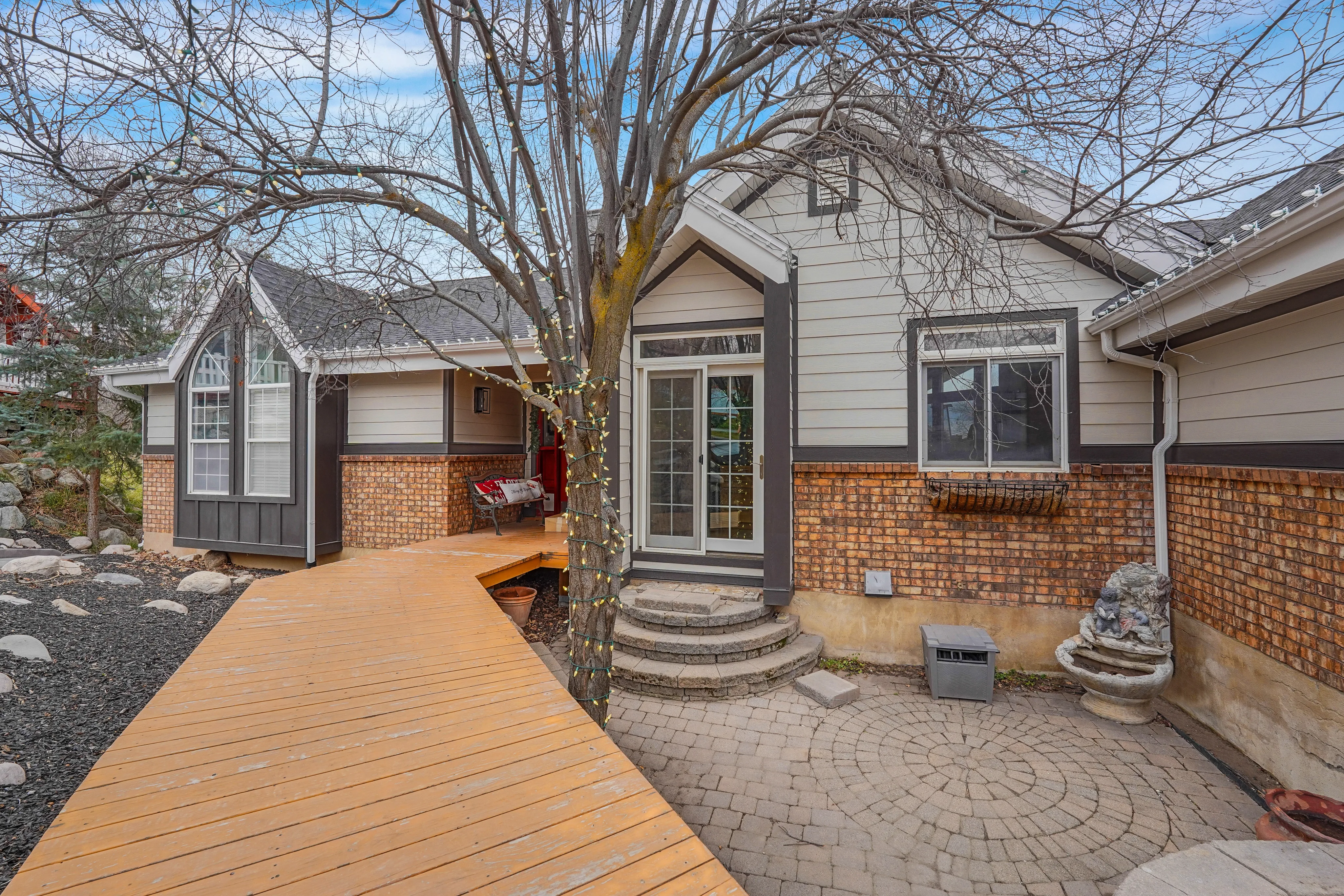 Front entry of a single-story home with brick siding, wood walkway, and stone patio with steps leading to glass doors
