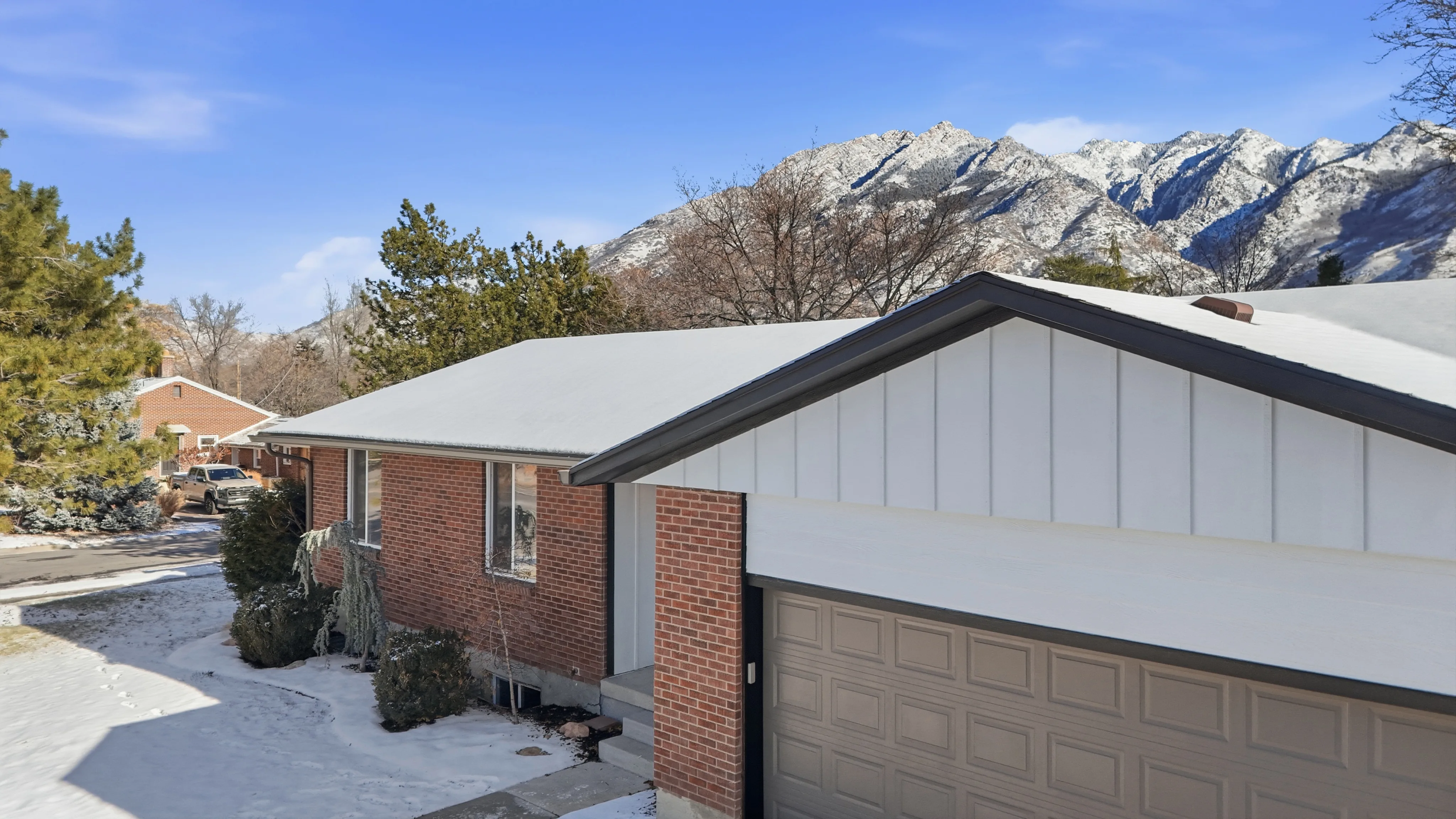 Brick home exterior with white board and batten siding, gray garage door, and snow-covered roof against Utah mountain backdrop